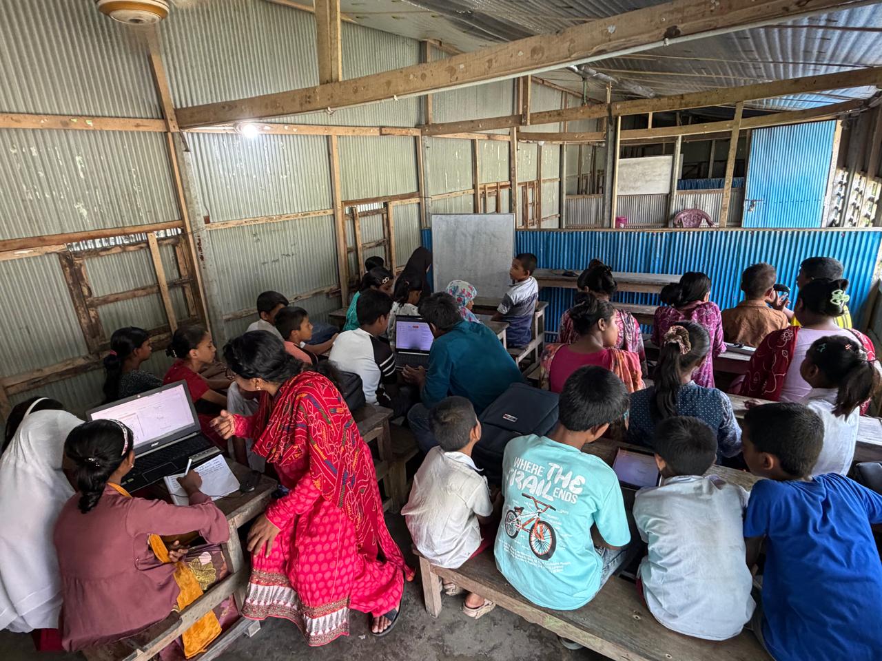 A full classroom of students with laptops in a tin-roofed school
